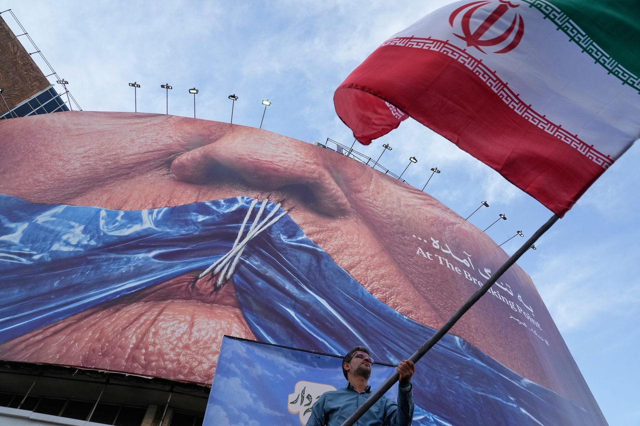 イラン、テヘラン市内の広場で男性がホルムズ海峡とドナルド・トランプ米大統領を描いた広告画像の前でイラン国旗を振っている。